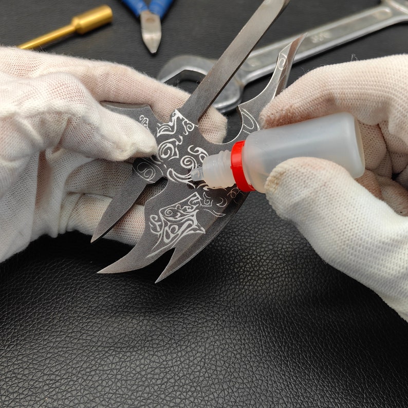 May include: A person wearing white gloves is applying clear glue to a metal object with a detailed design. The object has a sharp, pointed shape and appears to be a decorative piece.