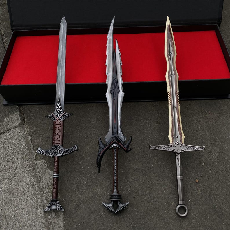 May include: Three decorative swords with ornate designs. The swords have silver blades and detailed handles in brown, black, and gold. They are displayed in front of a black box with a red interior.