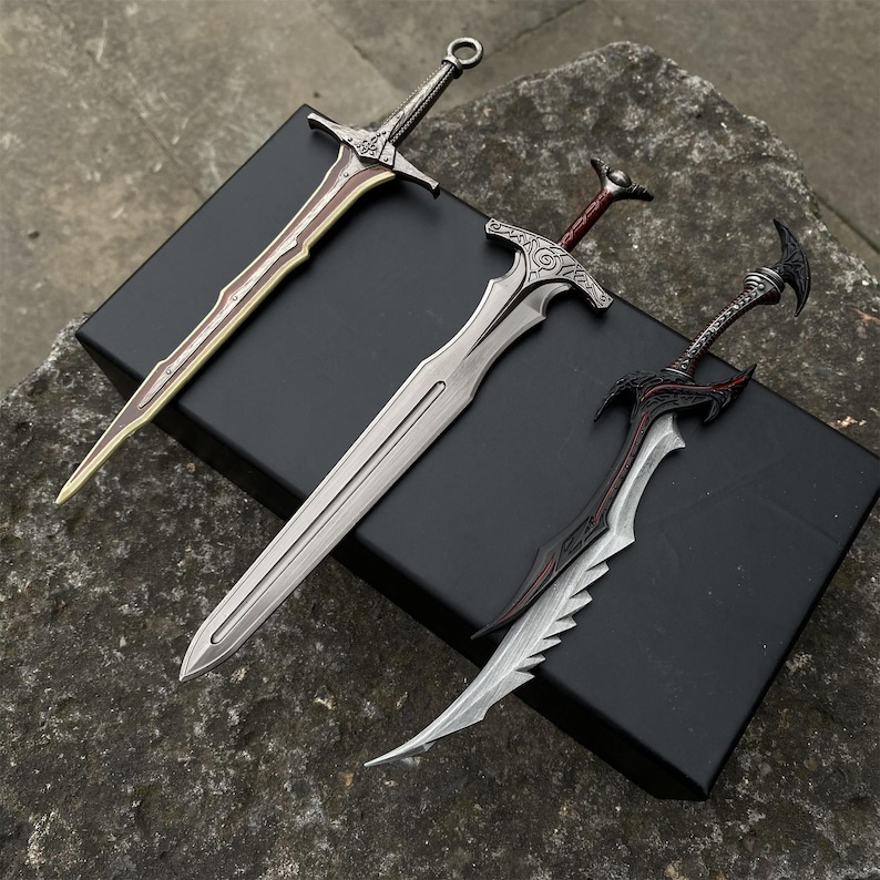 May include: Three decorative swords with intricate designs. The swords feature silver, gold, and black blades with detailed handles. The swords are displayed on a black box, set against a textured gray surface.