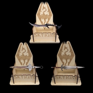 May include: Three wooden display stands, each holding a different miniature sword. The stands are light brown with the word "SKYRIM" and a dragon emblem. The swords vary in design, with silver and black blades.