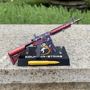 May include: A miniature rifle model featuring a red and black digital camouflage pattern, mounted on a black display stand labelled COUNTER-STRIKE. The stand includes a gold-coloured bullet replica and a circular logo depicting a white silhouette figure.