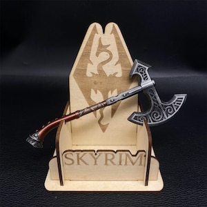May include: A miniature axe with a silver blade and a brown and silver handle rests in a wooden display stand. The stand is engraved with the word "SKYRIM" and a dragon emblem. The axe is a detailed replica.