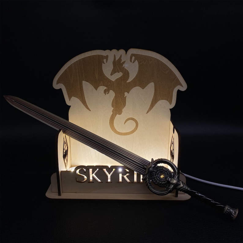 May include: A wooden Skyrim-themed night light featuring a sword and a dragon silhouette. The sword has a detailed hilt and blade. The base of the light has the word "SKYRIM" cut out. The light is powered by a cord.