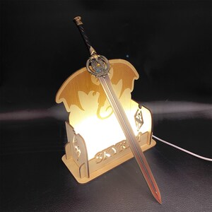 May include: A decorative sword with a detailed hilt and blade, resting on a wooden light-up stand. The stand features a dragon silhouette and the word "SKYR". The sword has a black handle and a red and silver blade.