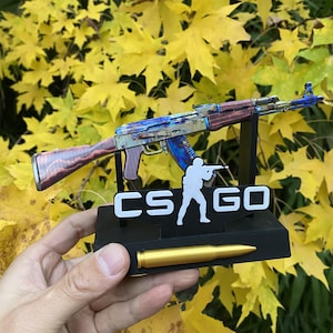 May include: A detailed model of an AK-47 assault rifle with a colourful, distressed finish. The model is mounted on a black base with the text "CS GO" and a soldier silhouette. A gold-coloured bullet is also included.
