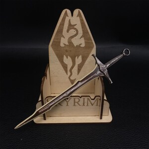 Puede incluir: Una espada detallada de color plateado con un mango marr&oacute;n descansa en un expositor de madera. El expositor presenta la palabra "SKYRIM" y un emblema de drag&oacute;n. La espada tiene una empu&ntilde;adura decorativa y una hoja afilada.