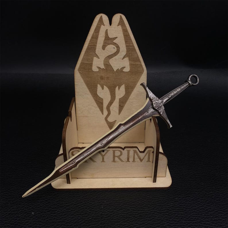 Puede incluir: Una espada detallada de color plateado con un mango marr&oacute;n descansa en un expositor de madera. El expositor presenta la palabra "SKYRIM" y un emblema de drag&oacute;n. La espada tiene una empu&ntilde;adura decorativa y una hoja afilada.