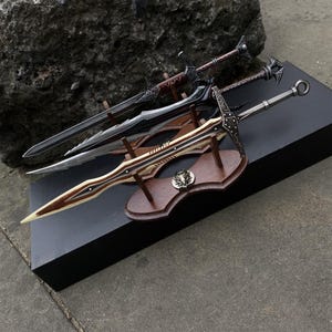 May include: Three decorative swords with ornate designs. The top sword has a dark brown handle and blade. The middle sword is black and silver. The bottom sword is brown and cream, mounted on a wooden display.