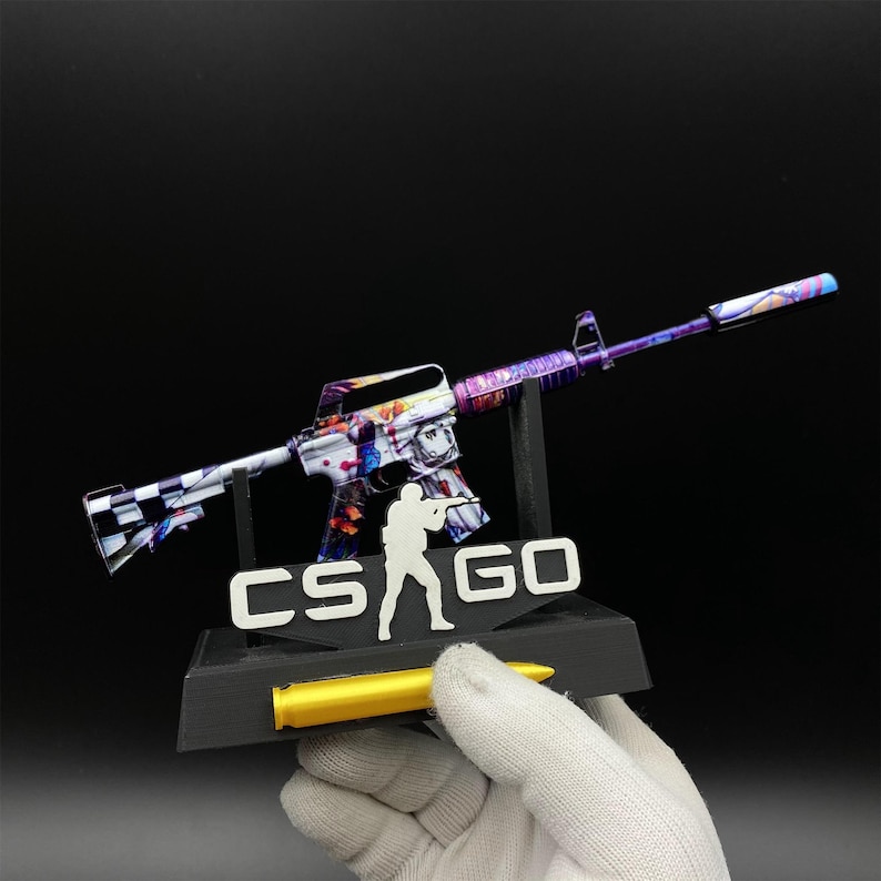 Pu&ograve; includere: Replica decorativa di un fucile con un design colorato, che include vernice rossa, blu e viola. La base mostra il testo "CS GO" e una silhouette di una persona. &Egrave; incluso anche un proiettile giallo.