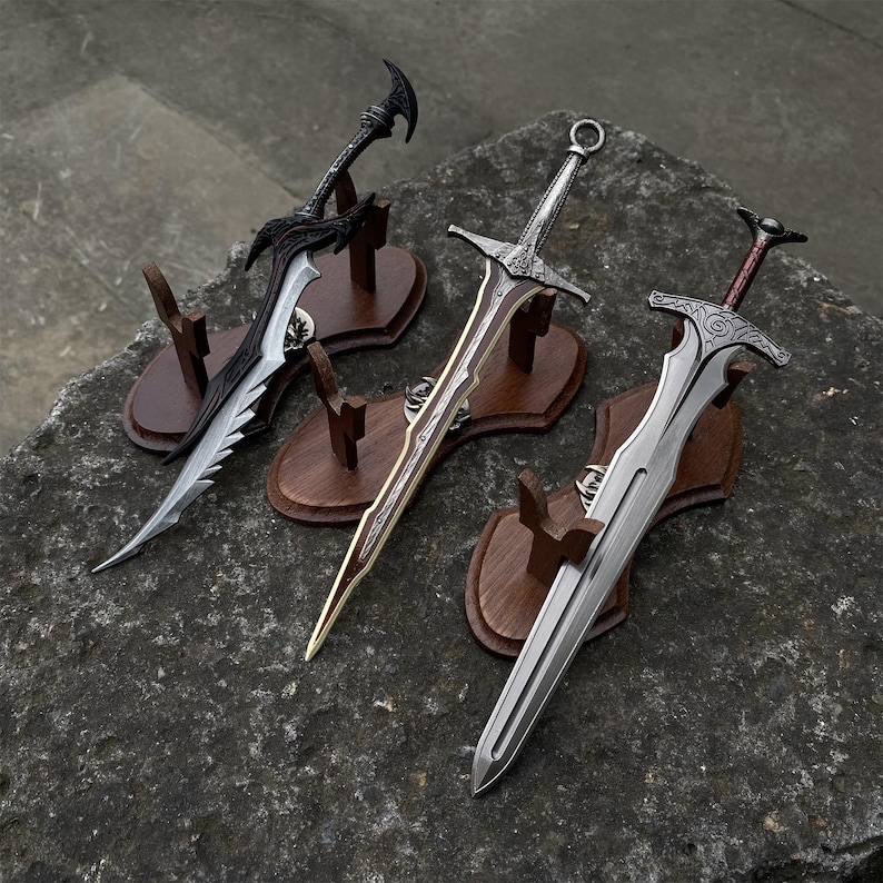 May include: Three decorative swords with ornate designs are displayed on wooden stands. The swords feature silver and black blades, with intricate hilt details. The stands are mounted on a textured, gray stone surface.