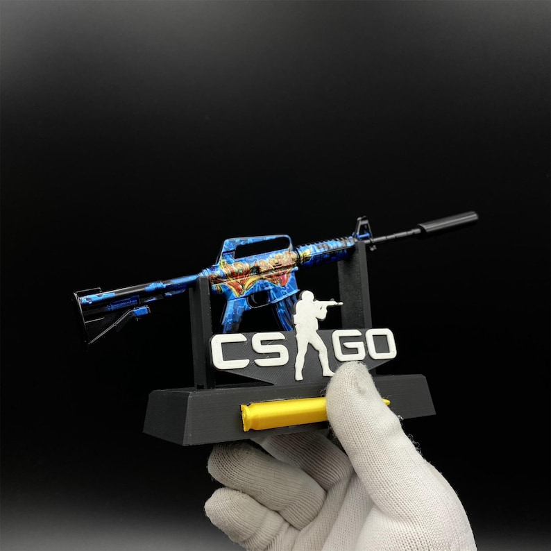 Pu&ograve; includere: Modello di fucile decorativo blu e oro con silenziatore, esposto su un supporto nero. Il supporto presenta il testo "CS GO" e un accento giallo. Il modello &egrave; tenuto da una mano guantata.
