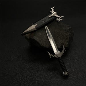 Puede incluir: Dos espadas met&aacute;licas en miniatura con detalles plateados ornamentados. Una espada est&aacute; desenvainada, mostrando una hoja afilada y un mango negro. La otra espada est&aacute; envainada en una vaina negra con detalles plateados. Las espadas se exhiben sobre una roca oscura.