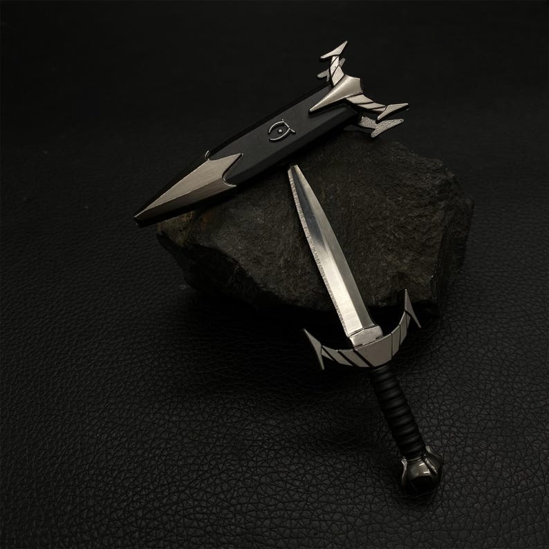 Puede incluir: Dos espadas met&aacute;licas en miniatura con detalles plateados ornamentados. Una espada est&aacute; desenvainada, mostrando una hoja afilada y un mango negro. La otra espada est&aacute; envainada en una vaina negra con detalles plateados. Las espadas se exhiben sobre una roca oscura.