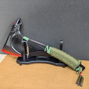 May include: Black axe with a crow head design, red accents, and a green paracord wrap. The axe is mounted on a black wooden stand.