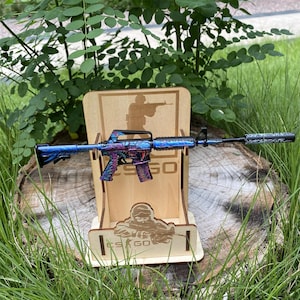 Op de afbeelding: Een decoratieve replica van een geweer met een blauwe, paarse en zwarte afwerking, gepresenteerd op een houten standaard. De standaard toont een silhouet van een persoon die een geweer vasthoudt en de tekst "CS GO".