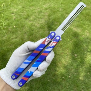 May include: A butterfly comb with a silver comb blade and blue handles. The handles have a red, white, and blue design with a face graphic. The comb is held against a green grassy background.