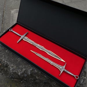 May include: Two decorative swords with silver handles and gold blades rest in a black box with a red velvet interior. The swords have intricate designs and are displayed at an angle, showcasing their detailed craftsmanship. The box is open, revealing the swords' full length.