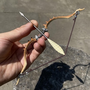 May include: A miniature bow and arrow set. The bow is a light brown color with dark brown thorns and silver accents. The arrow is silver with a cream-colored fletching. The bowstring is a dark brown cord.