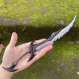 May include: A fantasy dagger with a silver blade and black handle, featuring red accents and intricate detailing. The blade has a serrated edge. The handle has ornate patterns and a curved pommel. The dagger is held in a hand.
