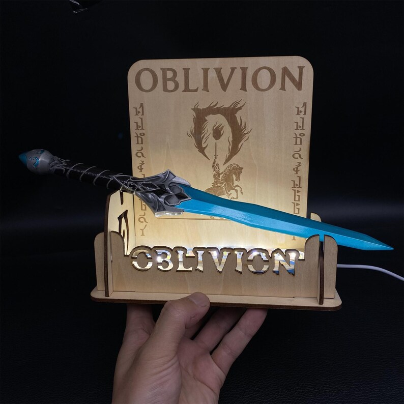 May include: A light-up wooden display featuring the word "OBLIVION" and a blue and silver sword. The display has a wooden back with the game's logo and additional text. The sword is held in place by the display.