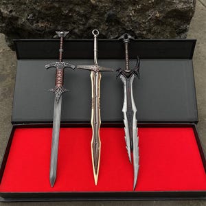 May include: Three decorative swords displayed in a black box with a red interior. The swords have ornate designs, with silver, brown, and black blades and handles. The swords are likely for display or cosplay.