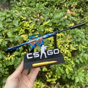 May include: A detailed miniature replica of a blue and black rifle with a silencer, resting on a black stand. The stand features the text "CS GO" and a gold-colored bullet. The rifle has a colorful design.