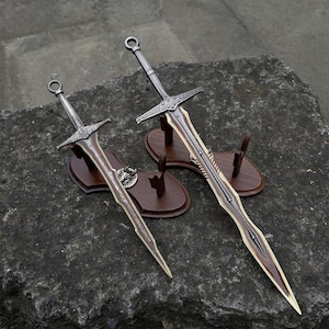 May include: Two decorative swords with ornate silver handles and brown blades are displayed on wooden stands. The swords have detailed crossguards and are set against a dark, textured background, showcasing their craftsmanship.