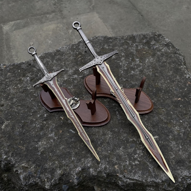 May include: Two decorative swords with ornate silver handles and brown blades are displayed on wooden stands. The swords have detailed crossguards and are set against a dark, textured background, showcasing their craftsmanship.