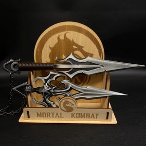 May include: Two silver-colored daggers with ornate designs and brown handles are displayed on a wooden stand. The stand features the text "MORTAL KOMBAT" and a dragon logo. A wooden disc with a dragon image is in the background.