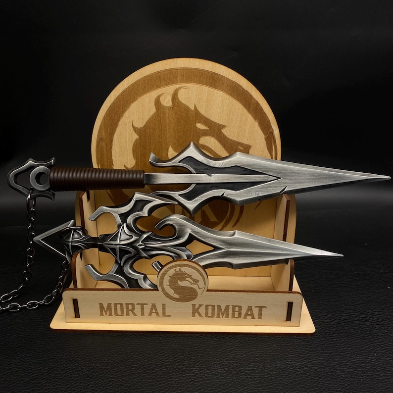 May include: Two silver-colored daggers with ornate designs and brown handles are displayed on a wooden stand. The stand features the text "MORTAL KOMBAT" and a dragon logo. A wooden disc with a dragon image is in the background.