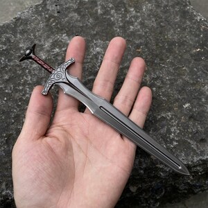 May include: A miniature sword with a silver blade and a decorative hilt. The hilt features an ornate design with a dark red grip and a black pommel. The sword is held in a hand against a gray stone background.