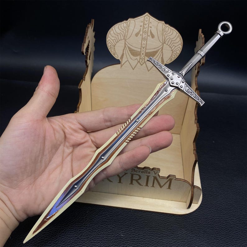 May include: A silver and bronze sword with a detailed hilt and blade, held in a hand. The sword is displayed in a wooden box with the text "Skyrim V" visible. The sword's blade has a blue and bronze design.