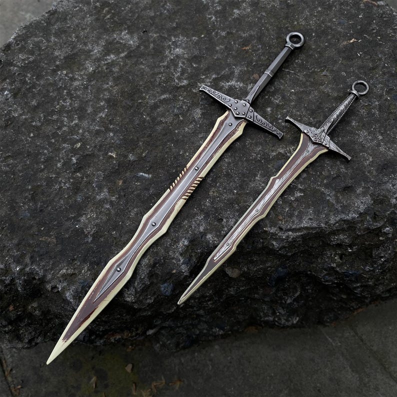 May include: Two decorative swords with ornate designs. The blades have a light brown center with a dark brown edge and a silver-colored spine. The hilts and crossguards are dark gray with decorative patterns. One sword is larger than the other.