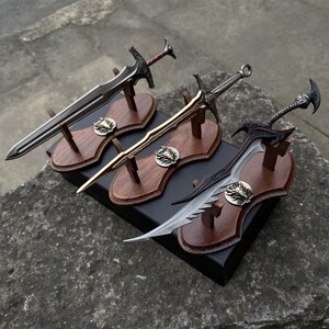 May include: Three decorative swords on wooden plaques. The swords have intricate designs and are displayed on dark wooden stands. Each plaque features a metal emblem. The swords vary in color, including silver, gold, and black.