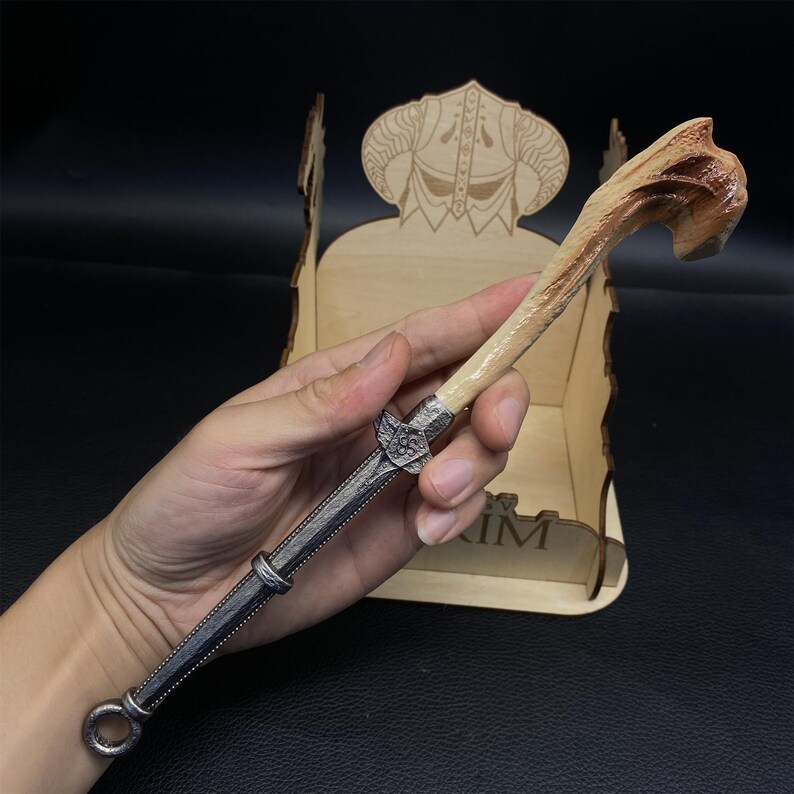 May include: A wand with a light brown, carved handle and a silver-colored metal shaft. The wand's handle has a textured, organic appearance, while the shaft features intricate detailing. The wand is held in front of a wooden display stand.