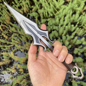 May include: A silver and black dagger with a pointed blade and ornate handle. The handle is wrapped in a brown, textured material. A small chain is attached to the end of the handle. The dagger is held in a hand against a green background.