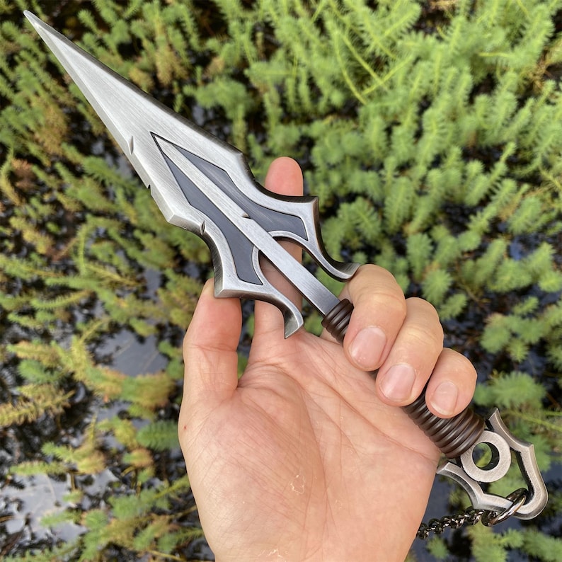 May include: A silver and black dagger with a pointed blade and ornate handle. The handle is wrapped in a brown, textured material. A small chain is attached to the end of the handle. The dagger is held in a hand against a green background.