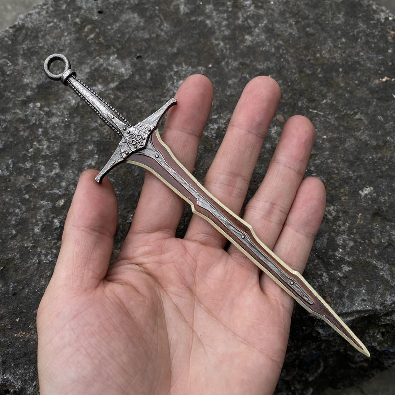May include: Miniature decorative sword with a silver-colored handle and crossguard, and a brown and cream-colored blade. The sword features intricate detailing on the handle and crossguard, and a ring at the top of the handle.