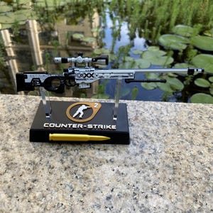 May include: A miniature sniper rifle model featuring a white and black patterned finish with a scope, mounted on a black display stand. The stand displays the text COUNTER-STRIKE, a circular logo with a figure, and a single gold-colored bullet replica.