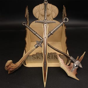 May include: A collection of miniature fantasy weapons, including a sword, axe, and two other bladed weapons, are displayed in a wooden box. The box has a decorative top with a horned design and the text "SKYRIM". The weapons have metallic and brown tones.