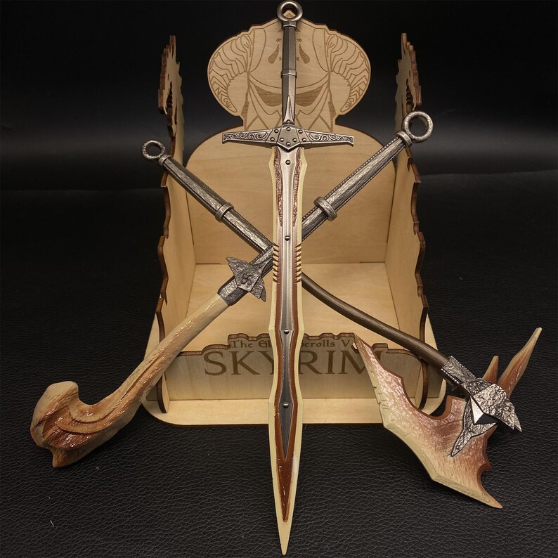 May include: A collection of miniature fantasy weapons, including a sword, axe, and two other bladed weapons, are displayed in a wooden box. The box has a decorative top with a horned design and the text "SKYRIM". The weapons have metallic and brown tones.