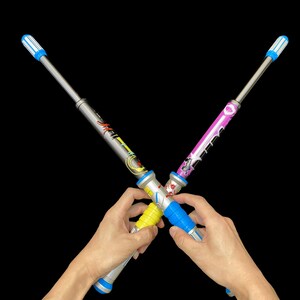 May include: Two toy light sabers are crossed against a black background. One saber is silver with a yellow and blue handle, and the other is pink with a blue handle. The tips are blue and white.