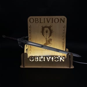 Könnte beinhalten: Ein Holzdisplay mit dem Wort "OBLIVION" und einem Schwert. Das Display ist hinterleuchtet und hebt das Schwert und das Logo des Spiels hervor. Das Schwert hat eine silberne Klinge und einen dunklen Griff.