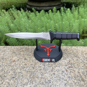 Puede incluir: Una daga plateada con mango negro descansa sobre un soporte negro con un emblema rojo y las palabras "RESIDENT EVIL". La daga tiene una hoja puntiaguda y un mango texturizado. El soporte est&aacute; sobre una superficie de piedra.