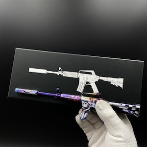 Pu&ograve; includere: Un oggetto decorativo a forma di fucile dai colori vivaci, presentato con una scatola nera con un contorno bianco di fucile. L'oggetto ha un design viola, blu e bianco. Tenuto da una mano guantata.