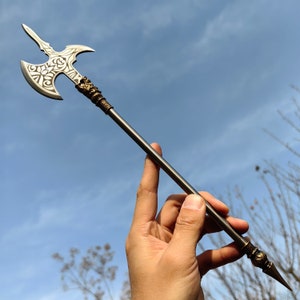 May include: A silver and bronze battle axe with a long, slender shaft. The axe head features a decorative floral design and two curved blades. The handle has ornate detailing near the head and the pointed end. The background is a blue sky.