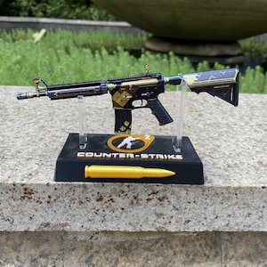May include: A miniature black and gold rifle replica mounted on a black display stand. The base features the text Counter-Strike, a circular logo with a figure, and a gold bullet. The rifle has intricate geometric patterns in gold.