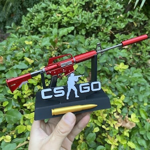 Puede incluir: Un rifle de juguete rojo con silenciador, exhibido sobre un soporte negro con el texto "CS GO" y una bala dorada. El fondo es un exuberante follaje verde. El rifle es una réplica de un arma de fuego real.
