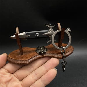 Puede incluir: Un llavero de espada en miniatura con una hoja negra y detalles plateados, exhibido en un soporte de madera. El soporte tambi&eacute;n presenta un peque&ntilde;o emblema met&aacute;lico y un llavero. El dise&ntilde;o es detallado y decorativo.