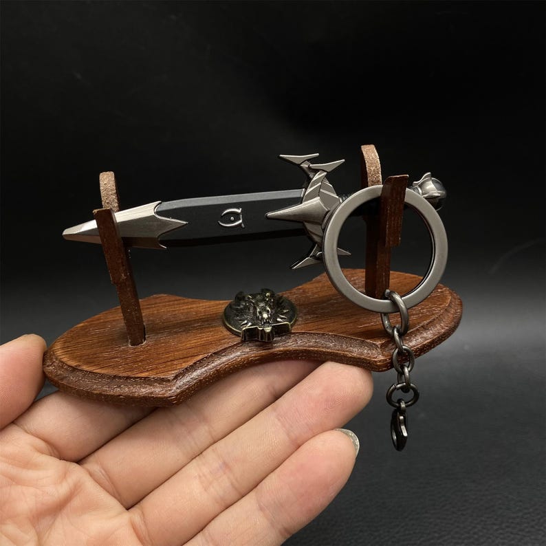 Puede incluir: Un llavero de espada en miniatura con una hoja negra y detalles plateados, exhibido en un soporte de madera. El soporte tambi&eacute;n presenta un peque&ntilde;o emblema met&aacute;lico y un llavero. El dise&ntilde;o es detallado y decorativo.
